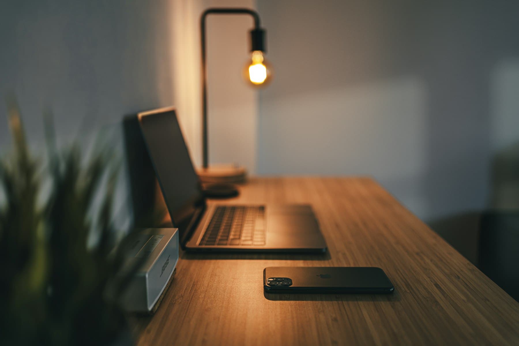 A dim wooden desk at night with a laptop, phone, and a warm desk lamp